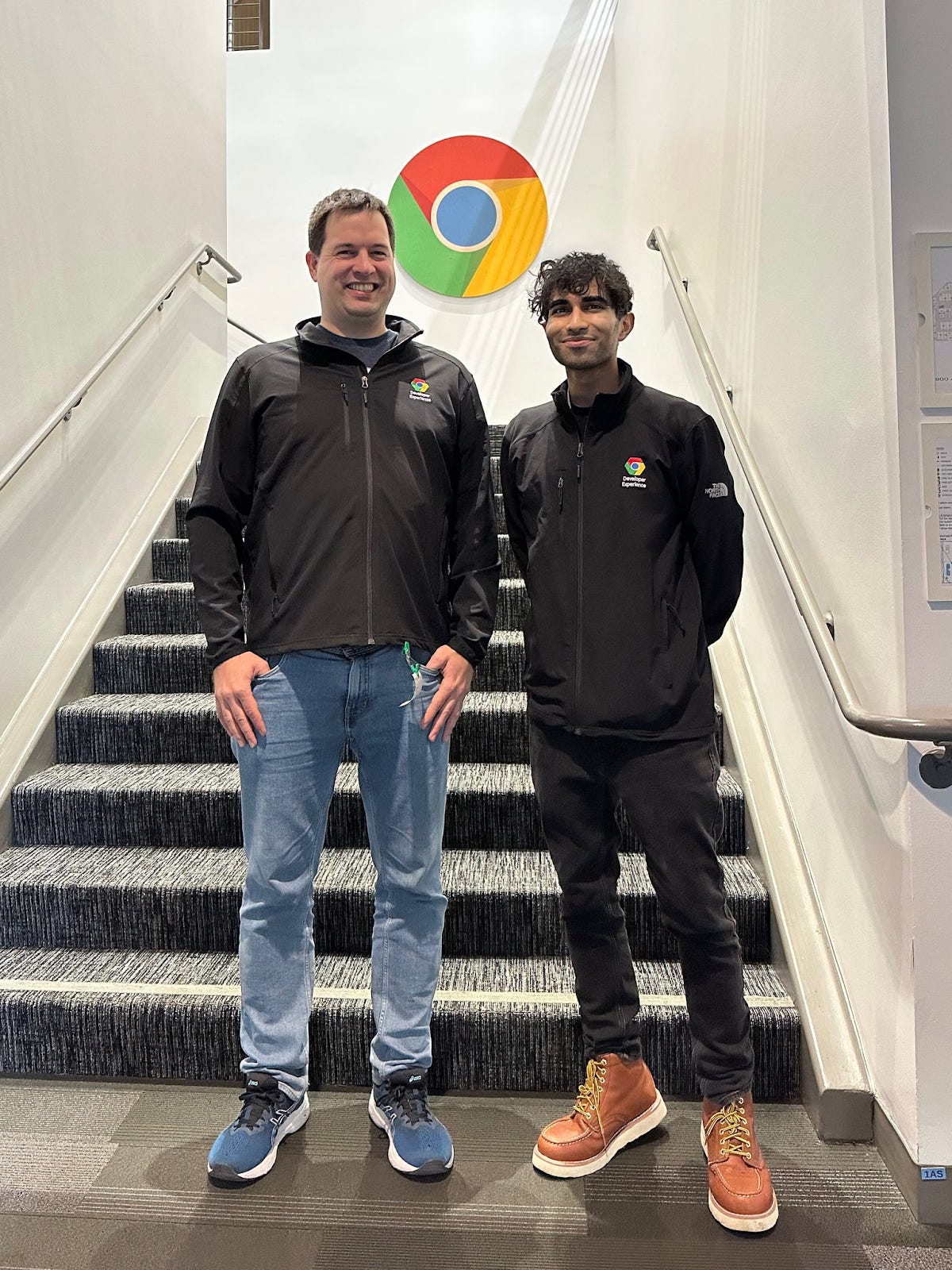 Two people standing on a staircase in a building with a large Chrome logo on the wall behind them. Both individuals wear Chrome-branded jackets and are smiling at the camera.